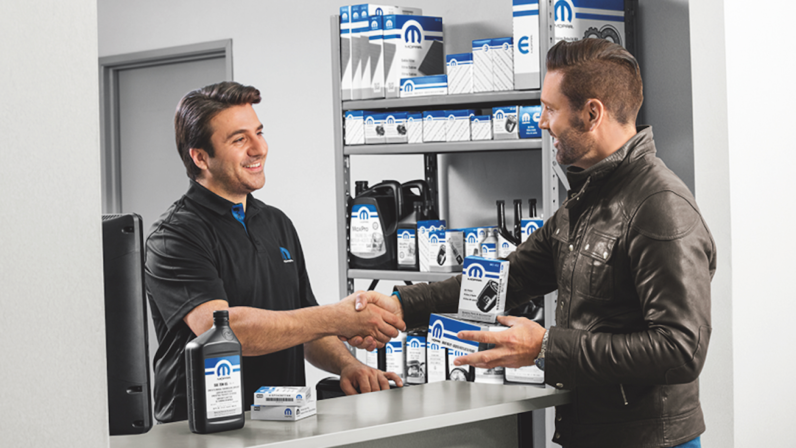 Chrysler, Dodge, Jeep or Ram dealer’s parts counter with a customer holding an oil filter and air filter in one hand, while shaking the dealer parts manager’s hand with the other, oil and spark plugs on the counter with other parts on a shelf in the background